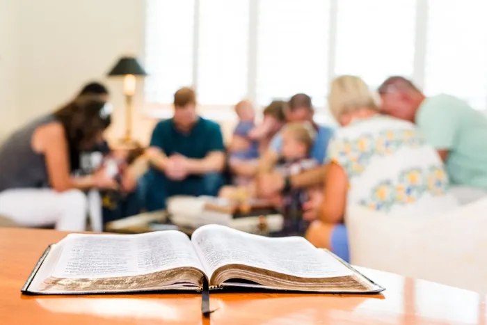 Bible lays open on a table in the foreground. An out-of-focus group of people pray in the background