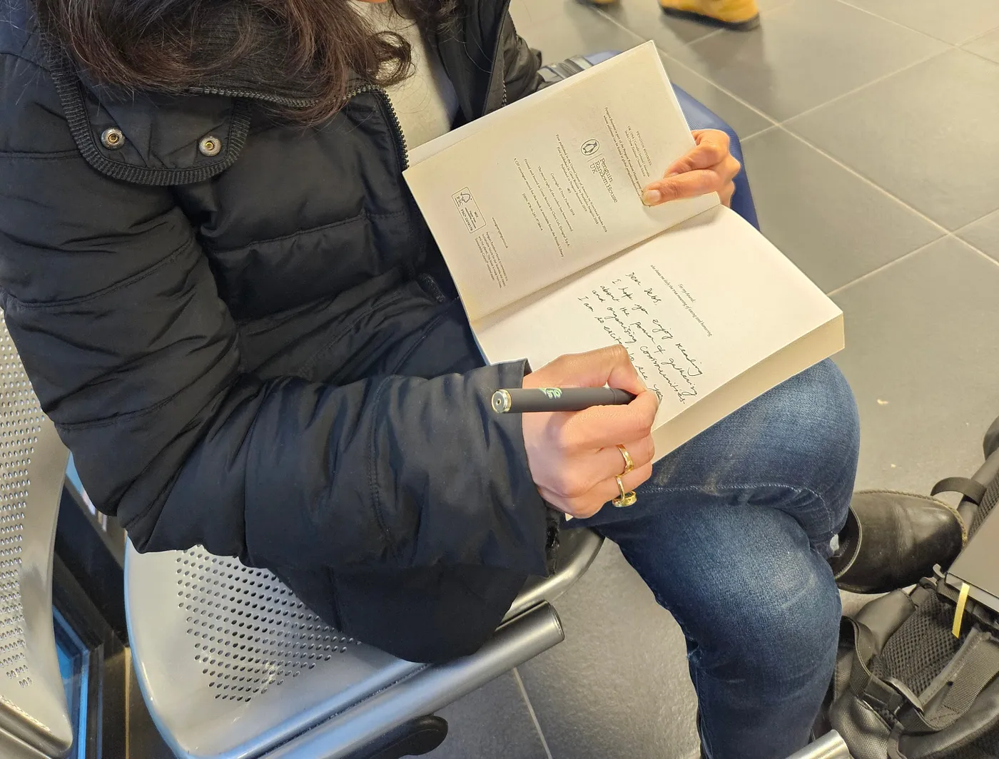 A woman wearing jeans and a black winter jacket sits on a metal chair, with her right leg crossed over the left. An open book is placed on her leg, and she is writing a note on the second preliminary page of the book, with a black pen.