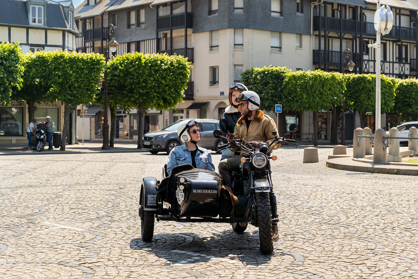 Three people wearing helmets sit on a vintage-style motorcycle with a sidecar, parked on a cobblestone plaza lined with neatly trimmed trees and European-style buildings in the background.