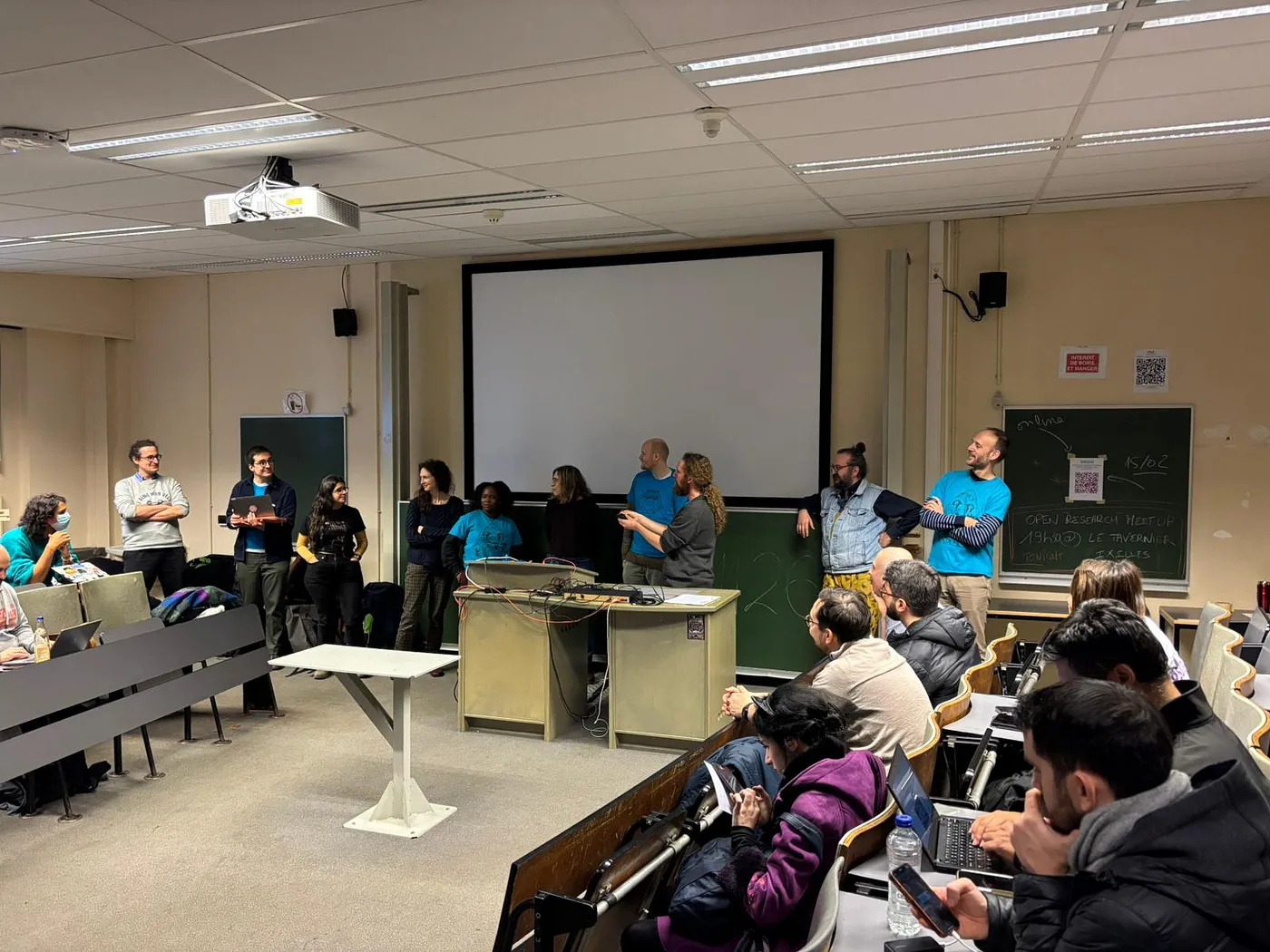 A small classroom, with attendees sitting in rows of chairs, forming an "n" shape. In front of the room is a group of ten (10) people from varying nationalities and gender groups. They are the members of the organising committee of the conference. Behind them is a blank projector screen, and in front, a beige table.