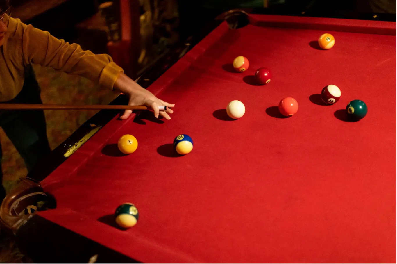 A person wearing a yellow long-sleeve shirt lines up a shot on a pool table with bright red felt. Their hand forms a bridge for the cue stick, aiming toward several scattered solid and striped billiard balls.