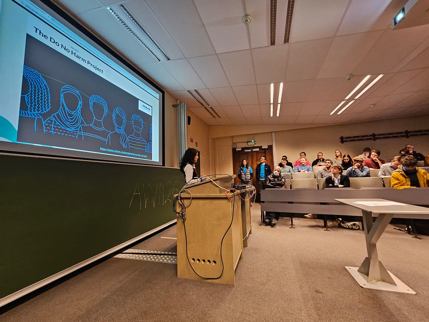 A conference room photographed from the left side of the room, showing a speaker, and some members of the audience. The speaker, Malvika, is an Indian woman of small stature and silky dark hair, wearing a white long-sleeved shirt under a sleeveless maroon dress. She is presenting from a slide that reads "The Do No Harm Project."