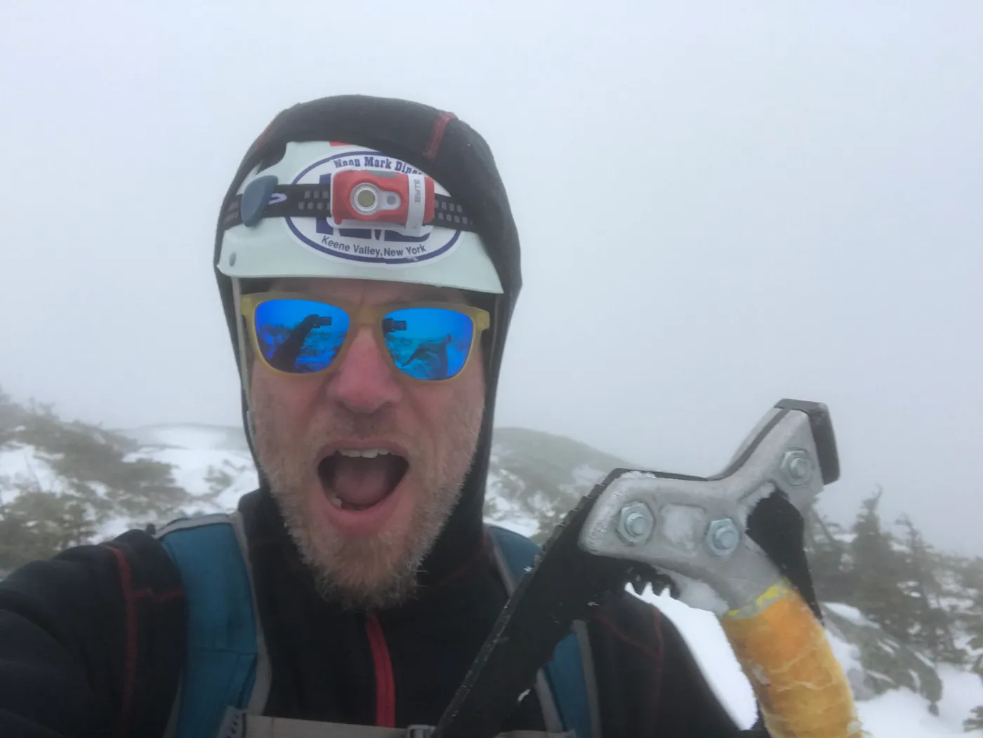 A mountaining stirring a hot drink over small gas stove with an ice climbing tool.