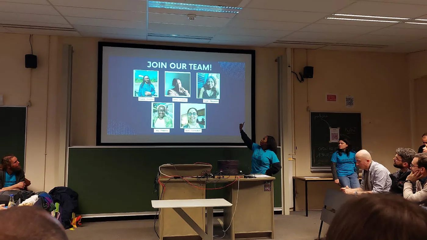 A conference room, with a speaker, Debs, delivering a talk. Debs is an African woman of average height and dark hair. She is wearing a blue "FOSDEM Organisers" t-shirt over her black long-sleeved sweater, and is pointing at the projected slide. The current slide is a blue page with a title in white text, which reads - "JOIN OUR TEAM" - with 5 members of an all-female team in display.