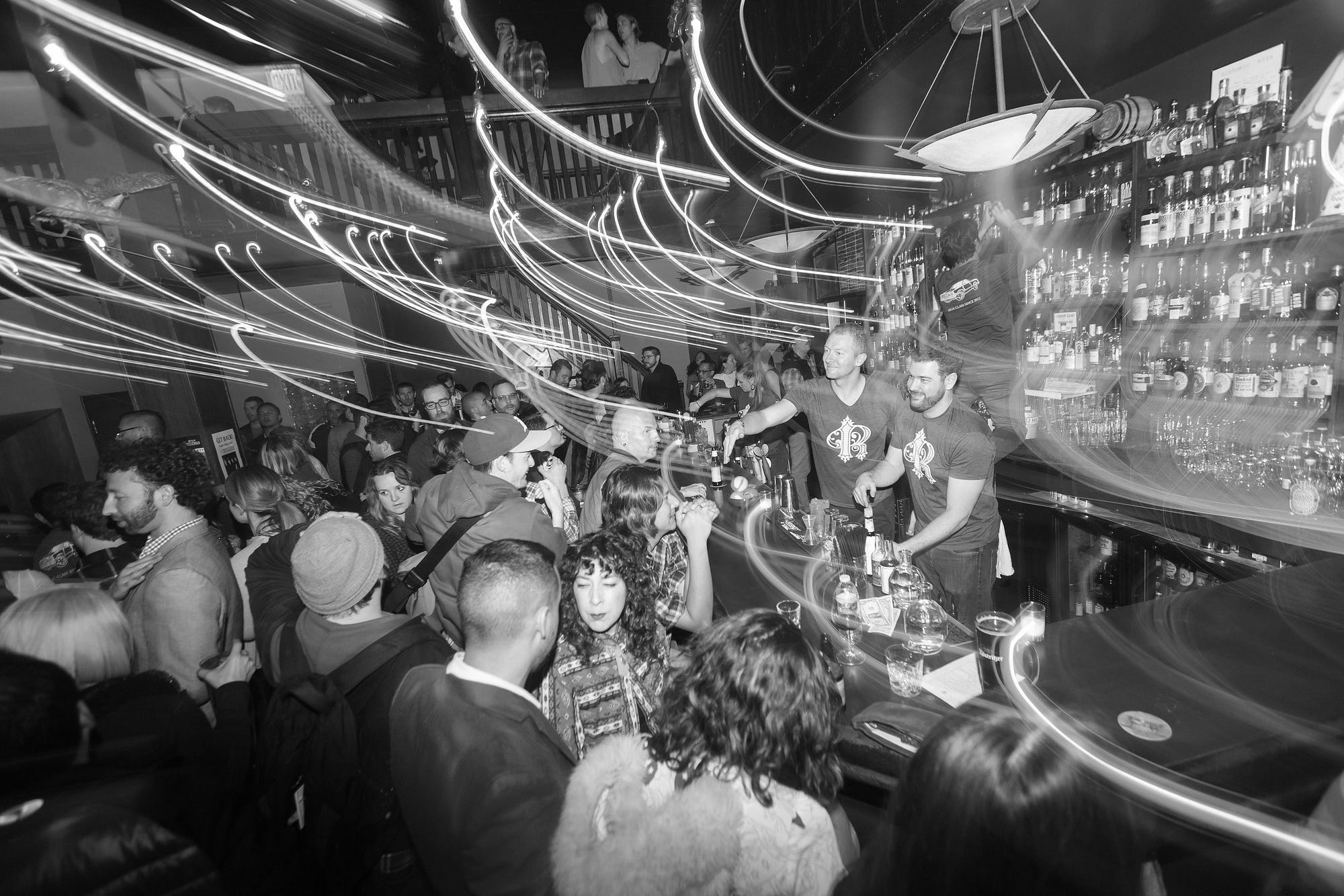 Long exposure black-and-white photo of a crowd of people around a bar with two bartenders.
