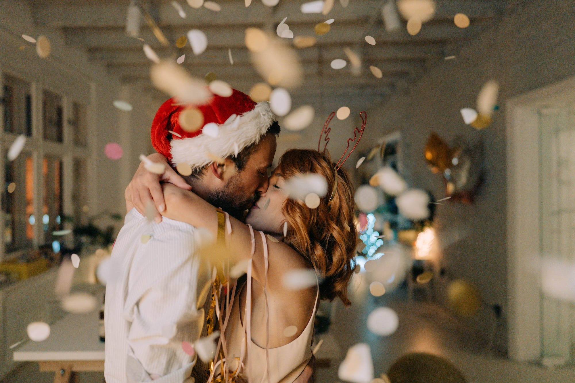 A young couple embrace and kiss with confetti fluttering around them. One has a Santa hat and the other, reindeer antlers.