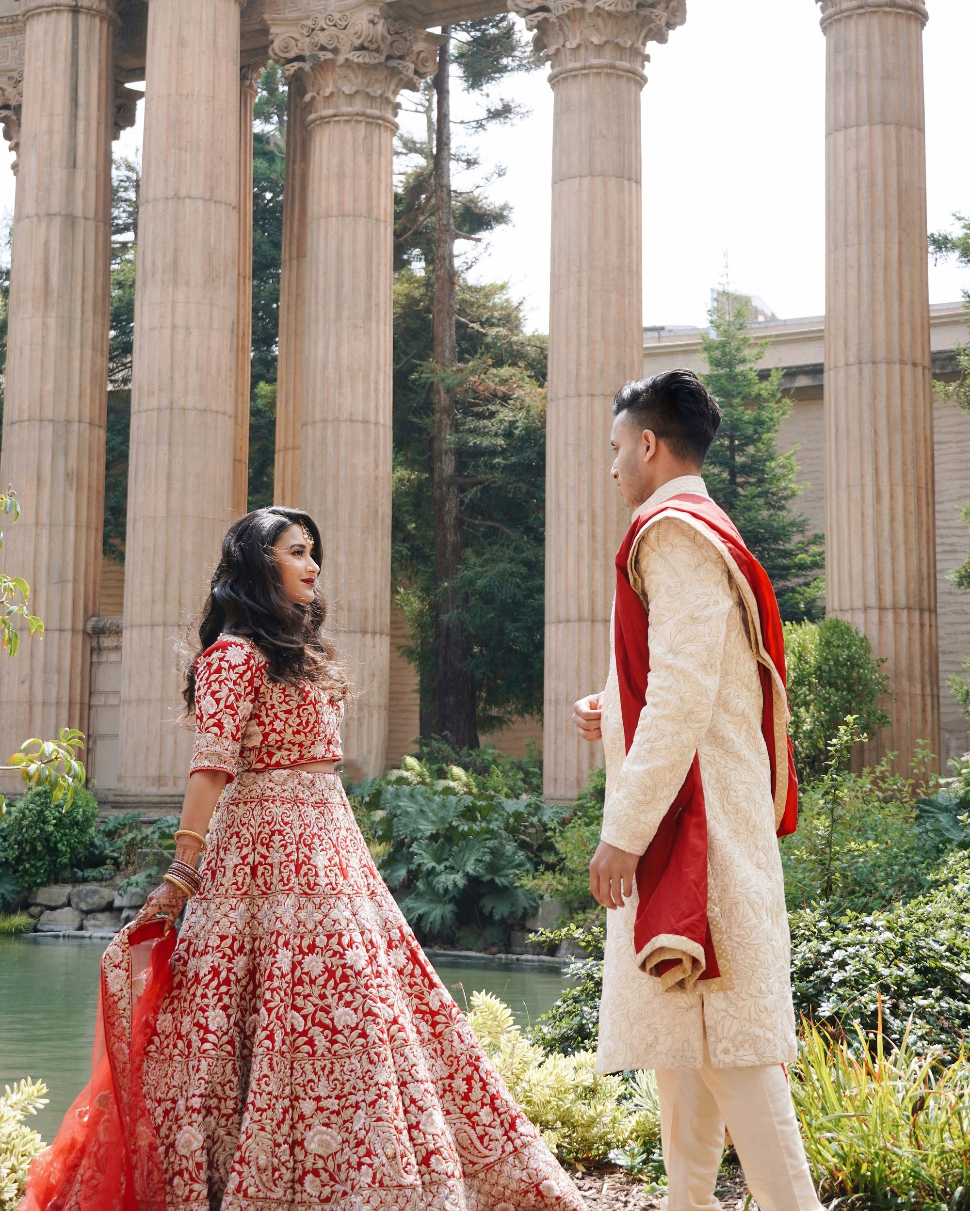 A bride and groom in traditional Indian wedding clothing standing in front of tall stone columns outside.