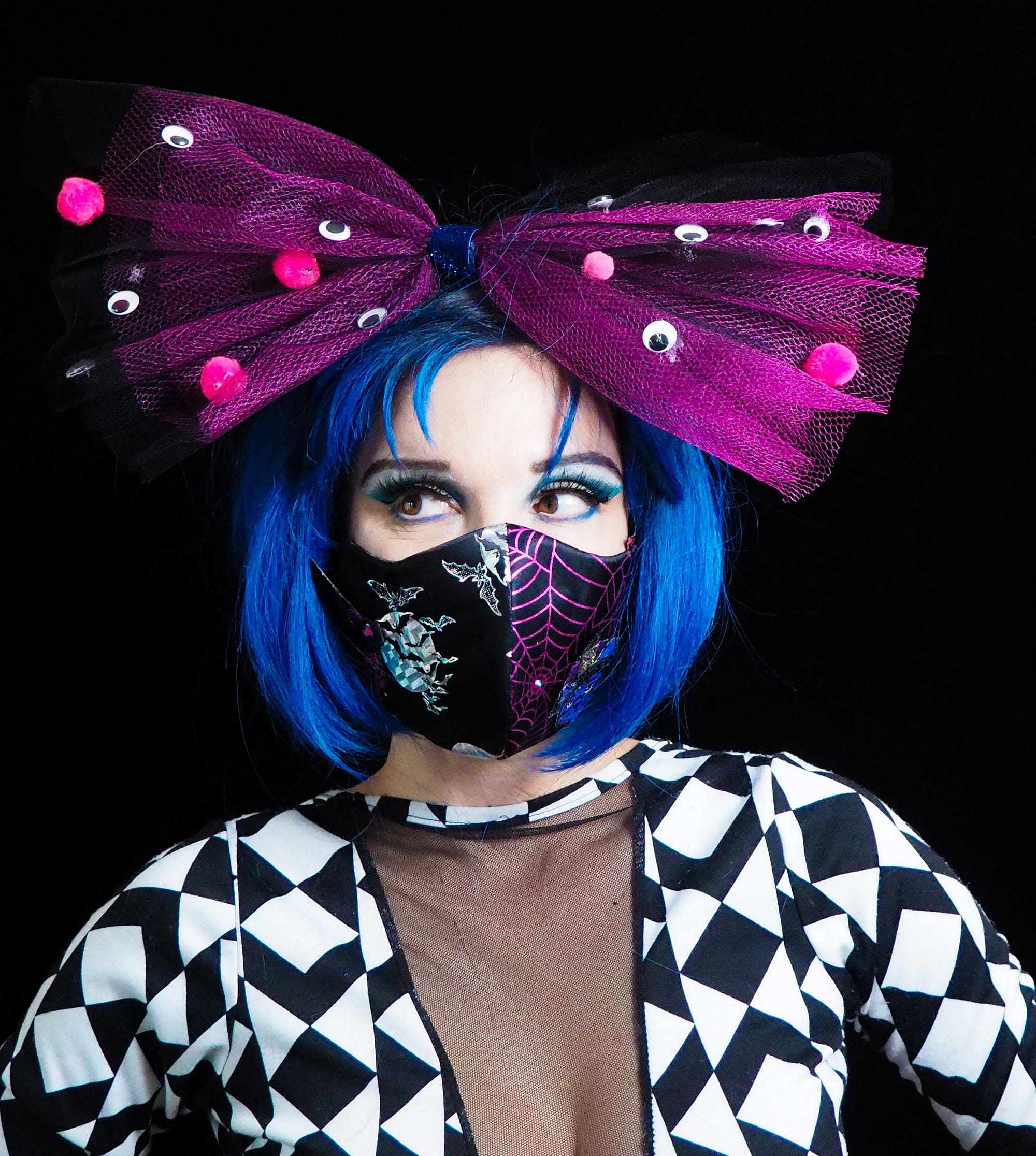 A person with a blue bob haircut wearing a huge purple hairbow and a mask with shiny bats + moons and purple spiderwebs