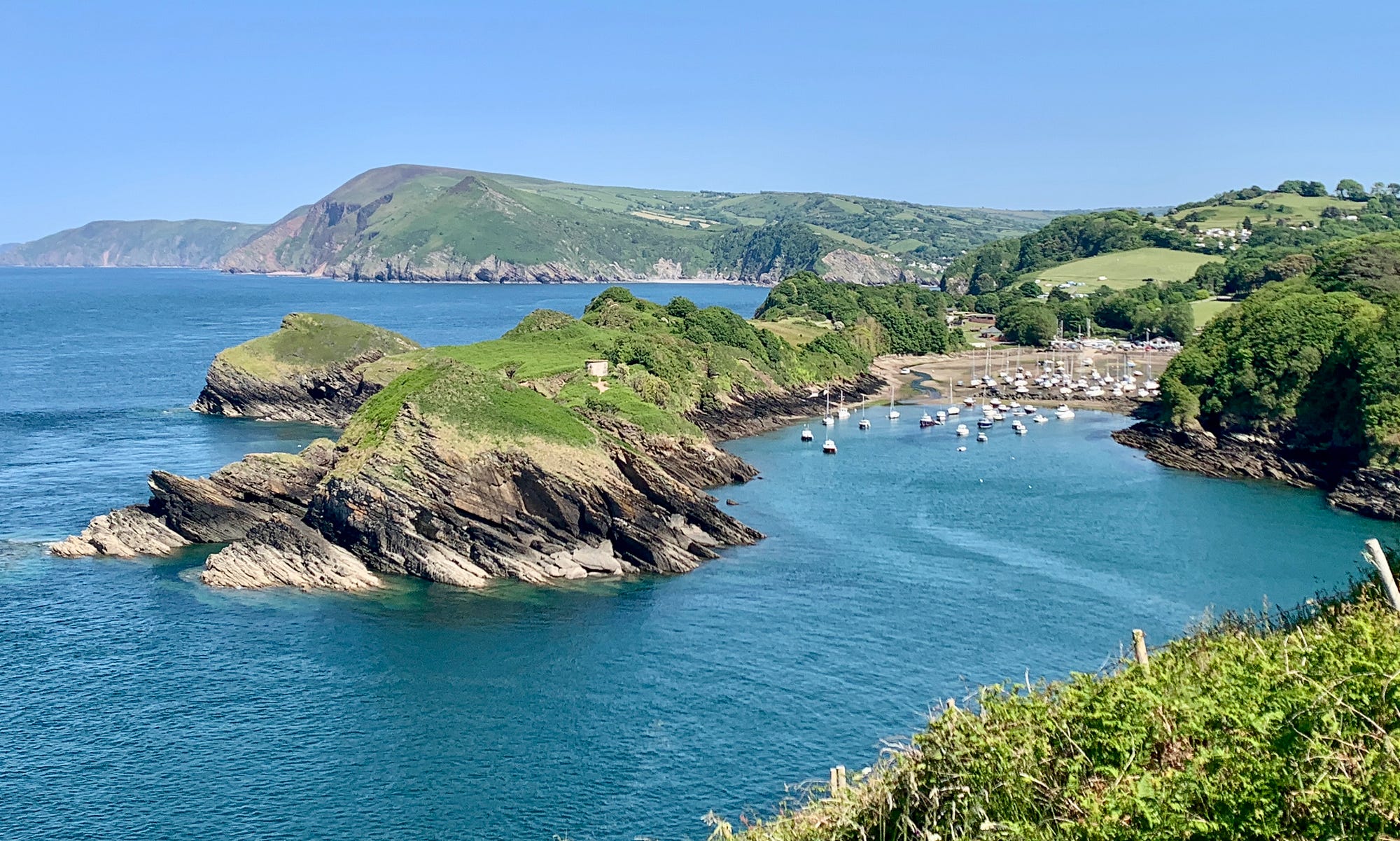 A coastal scene with blue water, a small cove with boats and grassy peninsulas.