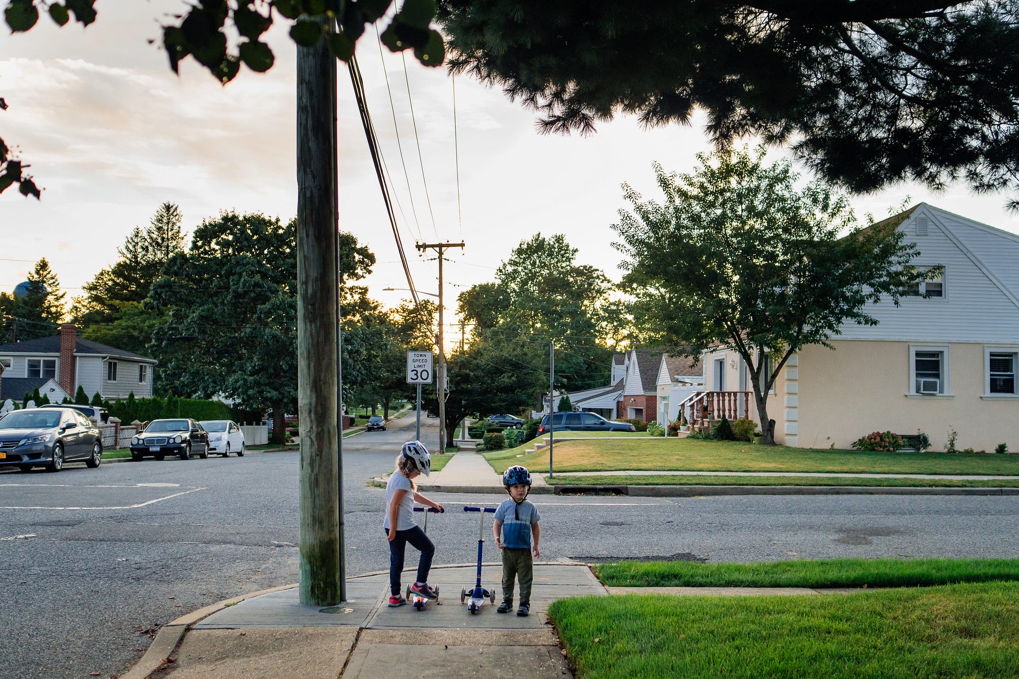 Two children stand on a residential street corner with their scooters.