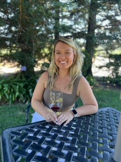 A women sits at a picnic table with a glass of red wine with evergreen trees in the background.