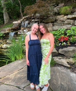 Two girls wear formal dresses with a rock wall and waterfall behind them.