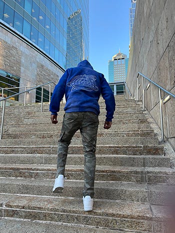A man in camo pants, blue hoodie and white sneakers walks up an urban staircase with his back turned to the viewer.