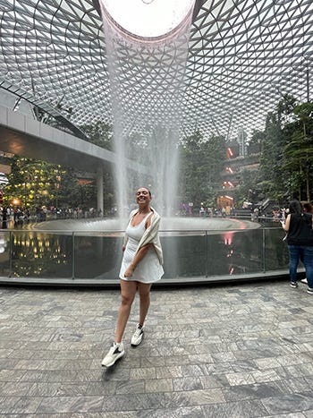 A girl in a white dress, sweatshirt and sneakers stands in front of an indoor waterfall with a modern steel structure above it and trees and botanics behind it.