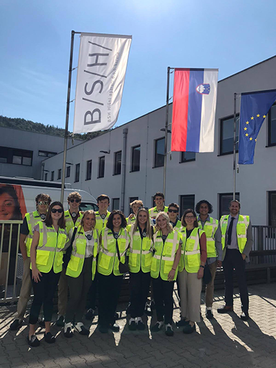 A group of students wearing yellow safety vests poses for a photo with three flags hanging above them depicting "B/S/H," the Slovenia flag and the E.U. flag.