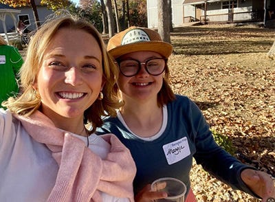 A girl with a pink sweater tied around her shoulders poses with a girl in a yellow hat with a nametag that reads "Maggie."