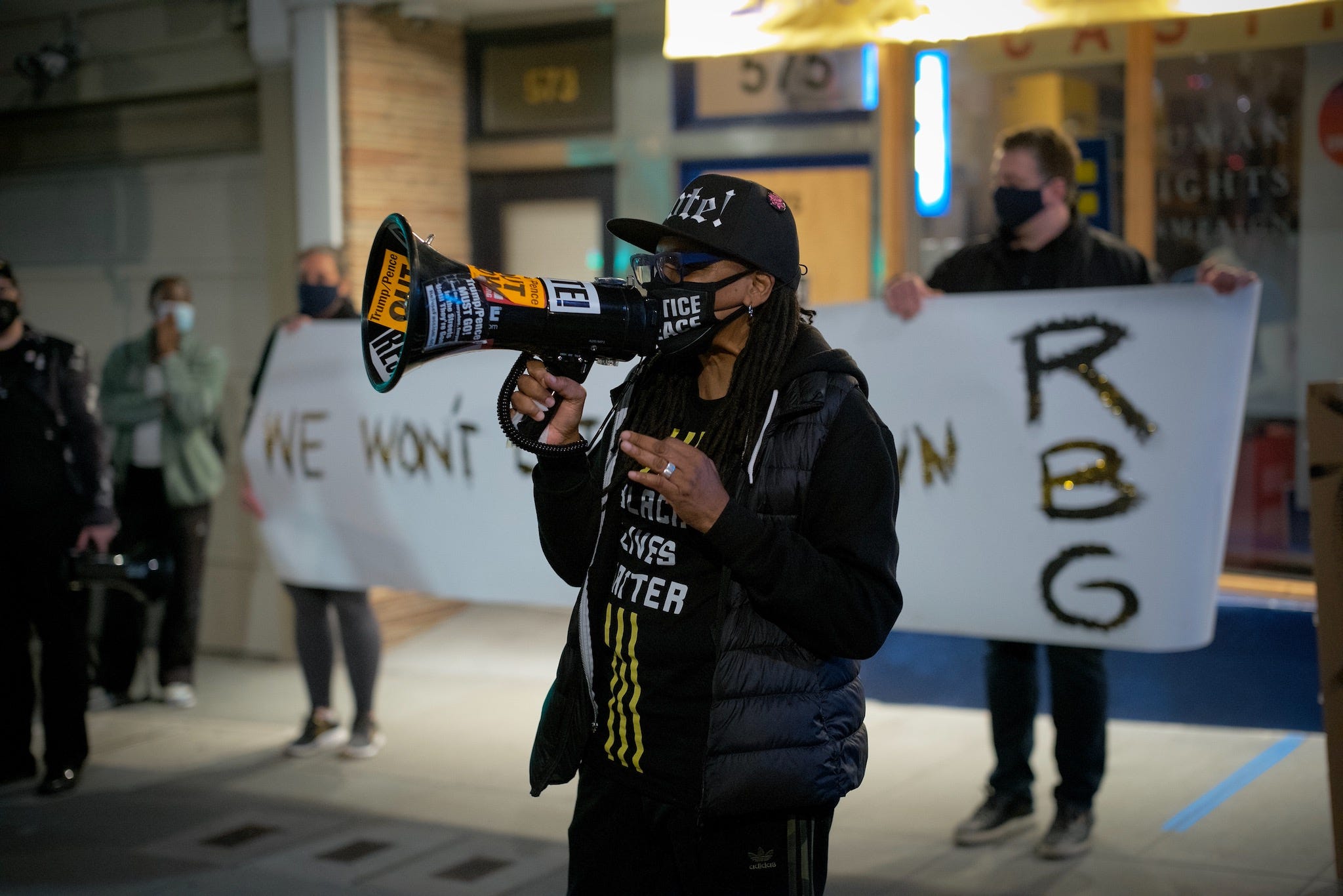 A person wearing a BLM shirt, hat, and mask, speaking into a megaphone in front of the white banner from the top photo.
