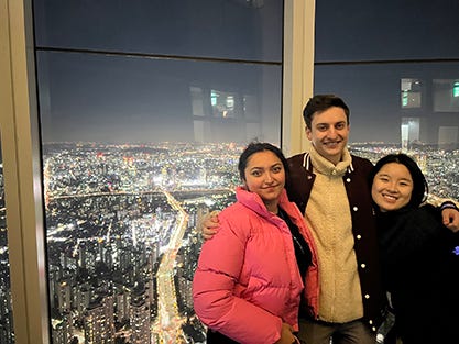 Three people pose for a photo against a glass wall overlooking an urban landscape at night.