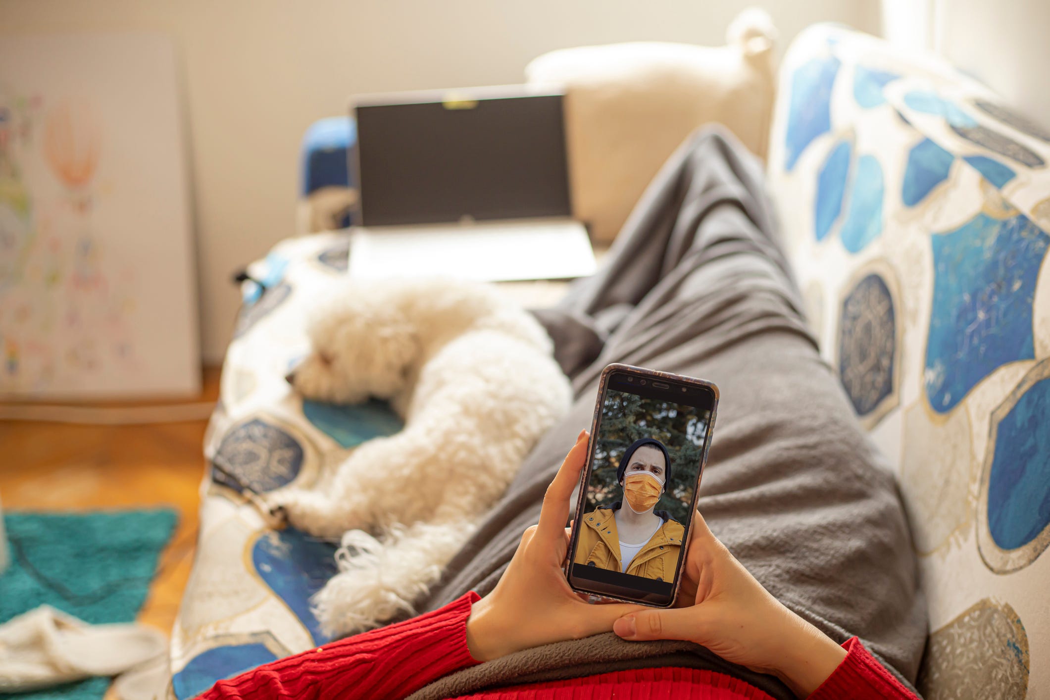 A person lying on a couch video holding a cellphone, video chatting with a masked person.