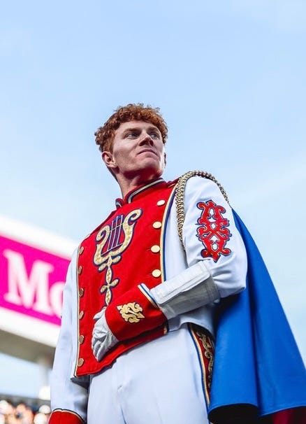 A man wearing a red, blue and white marching band drum major uniform stands with a clear sky in the background.