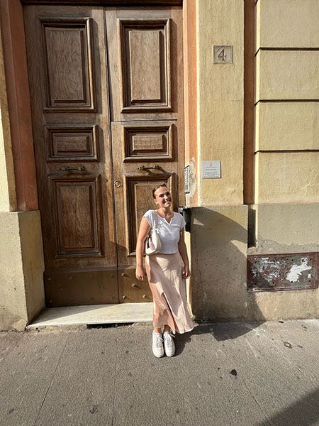 A girl stands in front of an older looking wood door dressed in a white shirt and pink skirt. 