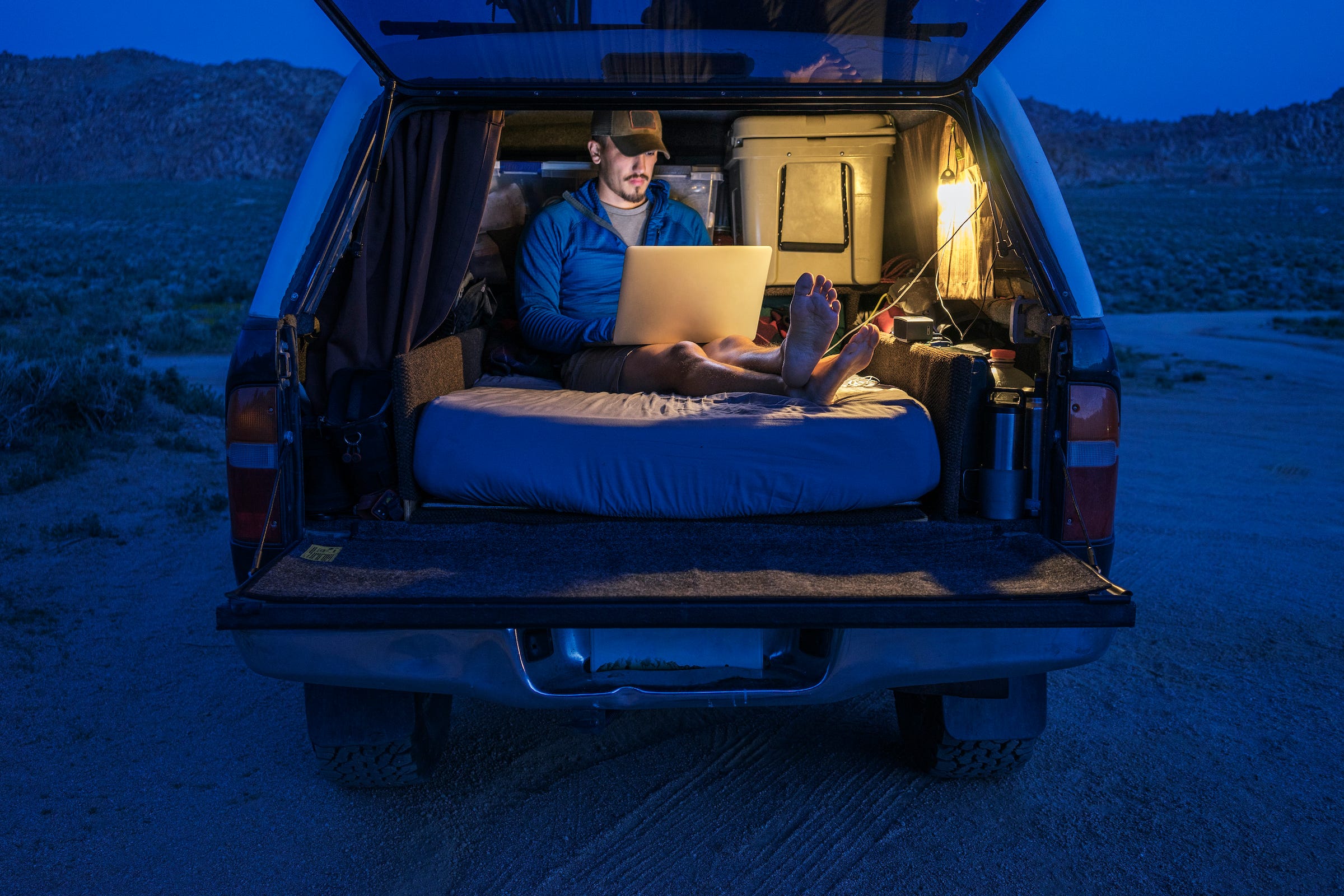 A person working on a laptop in the back of a covered pickup truck at night, sitting on a mattress.