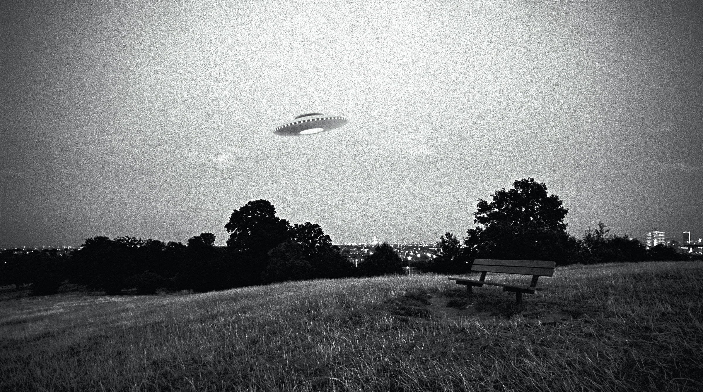 Grainy black-and-white photo of a UFO flying over a park bench overlooking an urban scene in the distance.