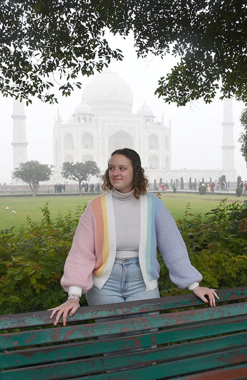 A girl wearing a pastel rainbow sweater stands behind a bench with a foggy Taj Mahal in the background. 