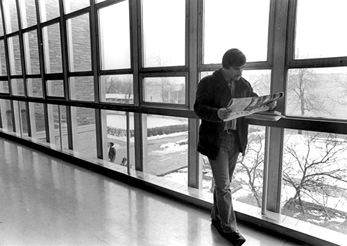 A black and white photo of a student reading a newspaper next to a wall of glass windows.