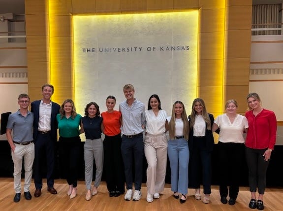 Students dressed in business casual attire pose for a photo in front of a lit up wall that says "The University of Kansas."