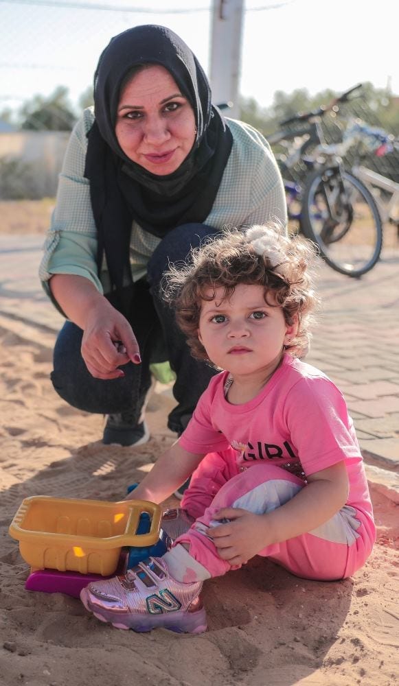 Wafaa and her niece Maryam at a MECA playground in Khuza’a, Gaza, in 2021. The photo shows a woman squatting by a child with a toy on a sandy playground with a bicycle in the background. Used with permission from MECA.