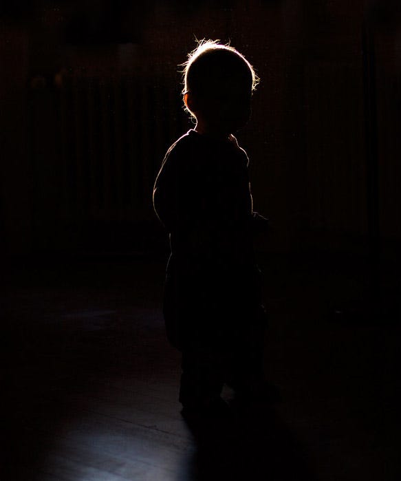 A young child stands alone in near-total darkness, seen only as a silhouette. A soft, warm light traces the outline of the child’s head and shoulders from behind, suggesting a halo or gentle glow. The child’s posture is still and quiet, facing slightly to the side. The surrounding darkness is deep and enveloping, yet the light gives the small figure a presence that feels both fragile and luminous.