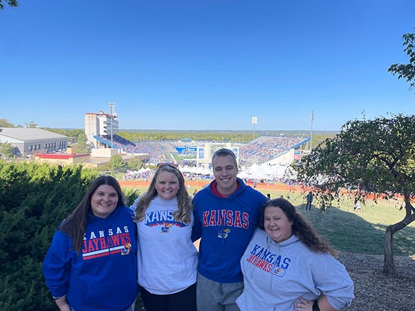 A group poses on a hill overlooking a football stadium while wearing KU sweatshirts.