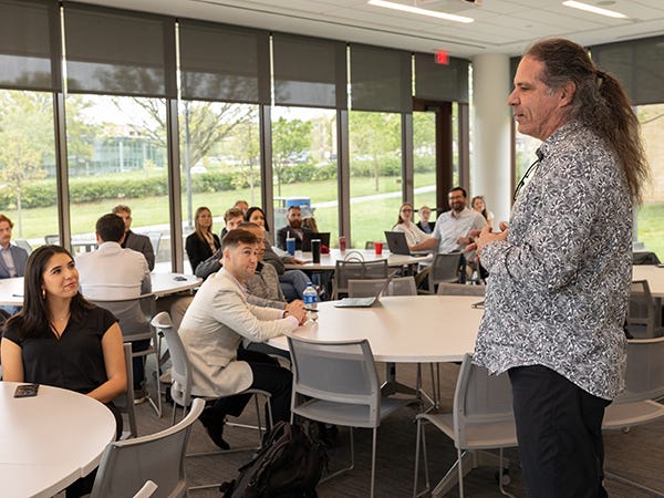 A man stands and speaks to people seated at round tables in a large classroom setting.