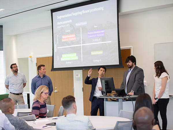 Students dressed in business attire present their projects on an overhead projector while attendees at roundtables look on.