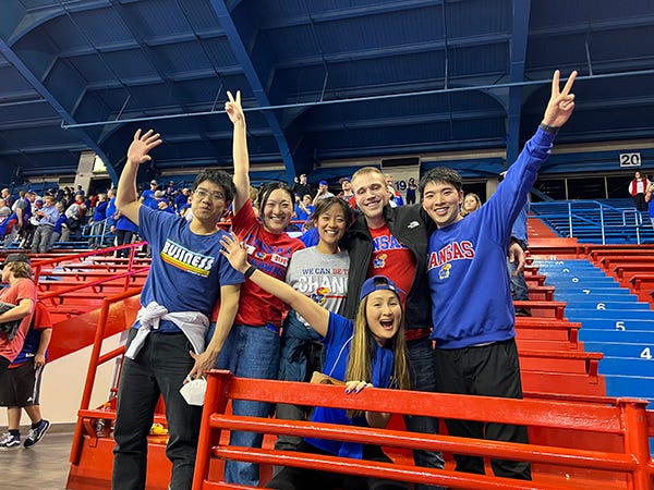 A group of students wearing red and blue KU fanwear pose at a basketball game on red and blue bleachers.