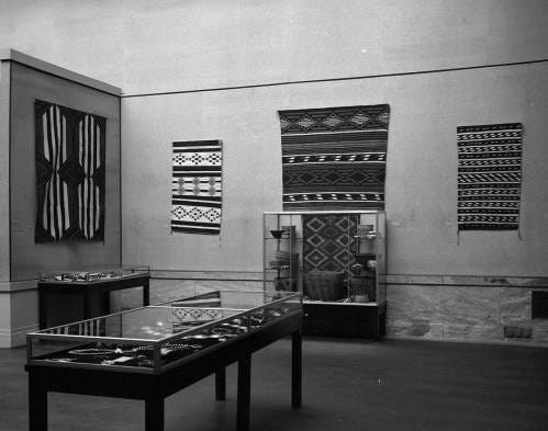 Installation image with textiles hanging on the walls and glass cases with objects.