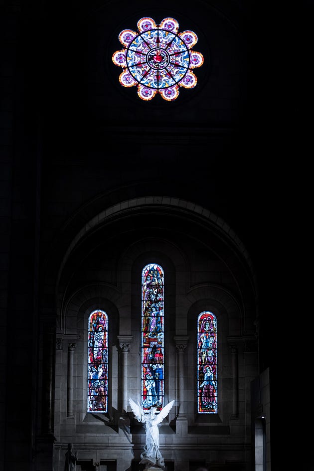 Stain glass windows in a church with a dark background and a beautiful statue of an angel in front.