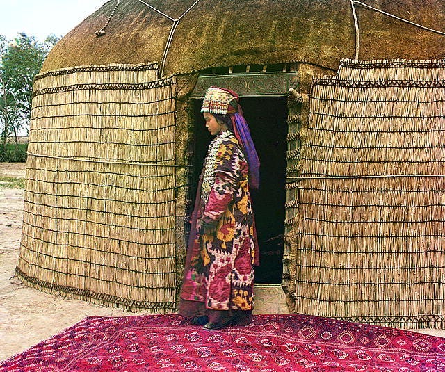 Full-length profile portrait of a woman, possibly Turkman or Kirghiz, standing on a carpet at the entrance to a yurt, dressed in traditional clothing and jewelry.