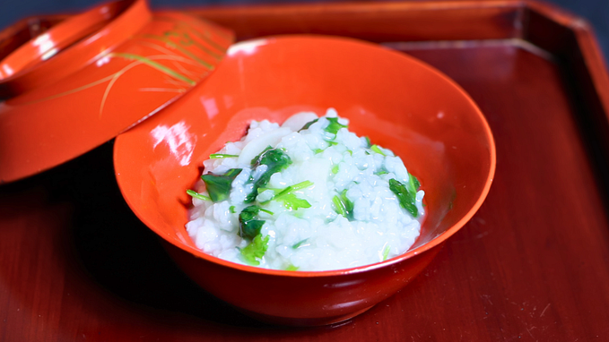 A traditional Japanese dish, nanakusa-gayu (seven-herb rice porridge), served in a bright orange lacquered bowl with a matching lid slightly ajar, placed on a wooden tray. The dish is garnished with fresh green herbs, symbolizing a refreshing and light New Year’s meal.