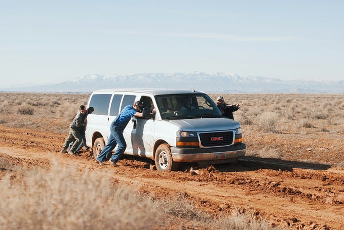 A group of people pushing a van stuck in the mud forward, with one person pointing to where they need to go.