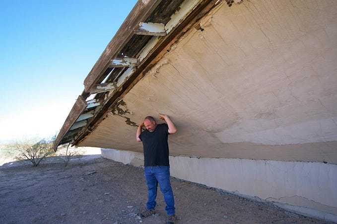 A person in black shirt and jeans standing under the foundation of a wooden house, seemingly keeping it from collapsing with his bare hands.
