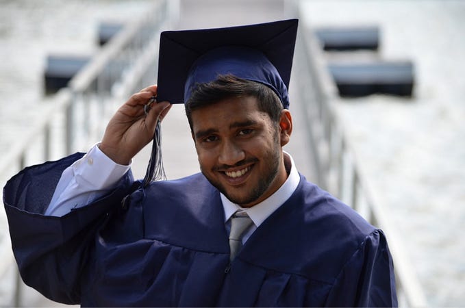A smiling, male graduate holding the tip of his mortarboard.