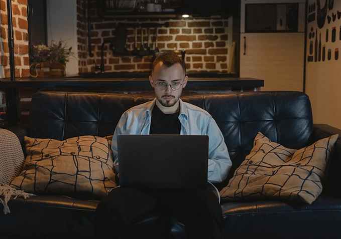 A photo of a young man working on a laptop computer in the family room in his house.