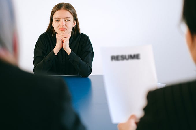 A woman with her elbows on the table and hands under her head, looking pensively at another person holding a piece of paper that says Resume