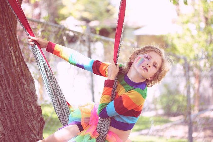 Image of a child on a swing, wearing a rainbow shirt, tutu and rainbow make-up