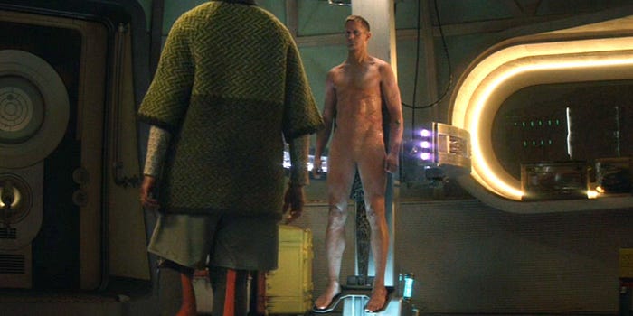 A naked android sans genitals stands in a repair bay while a woman looks on