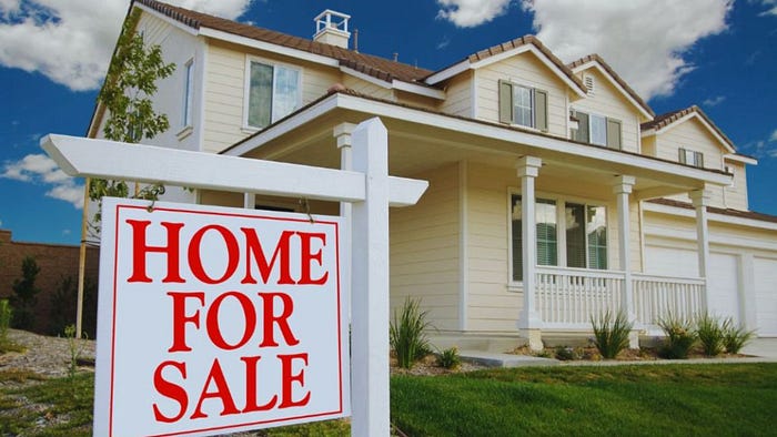 A large, two-story light beige suburban house with a front porch, white trim, and a manicured lawn is shown under a blue sky with scattered clouds. In the foreground, a white post displays a bright "HOME FOR SALE" sign in red letters.
