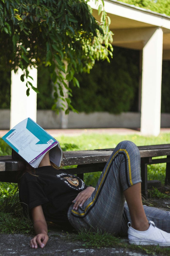 A woman leaning her head on a bench with her book on her face, showing signs of boredom