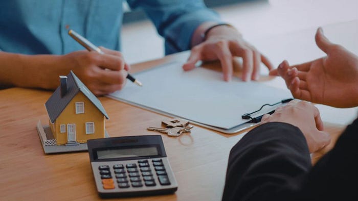 Real estate agent and client sitting at a desk reviewing a contract, with a small model house, calculator, and house keys in the foreground.
